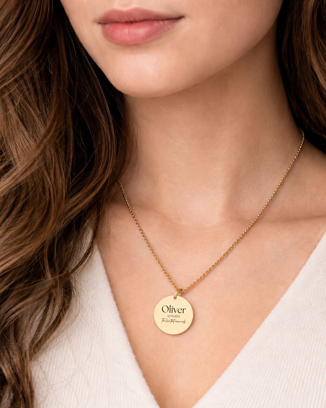 Close-up of a woman wearing a gold round pendant necklace with the name “Oliver,” a date, and a small handwritten-style message engraved on it. The necklace is styled with a cream V-neck sweater for a soft, elegant look.